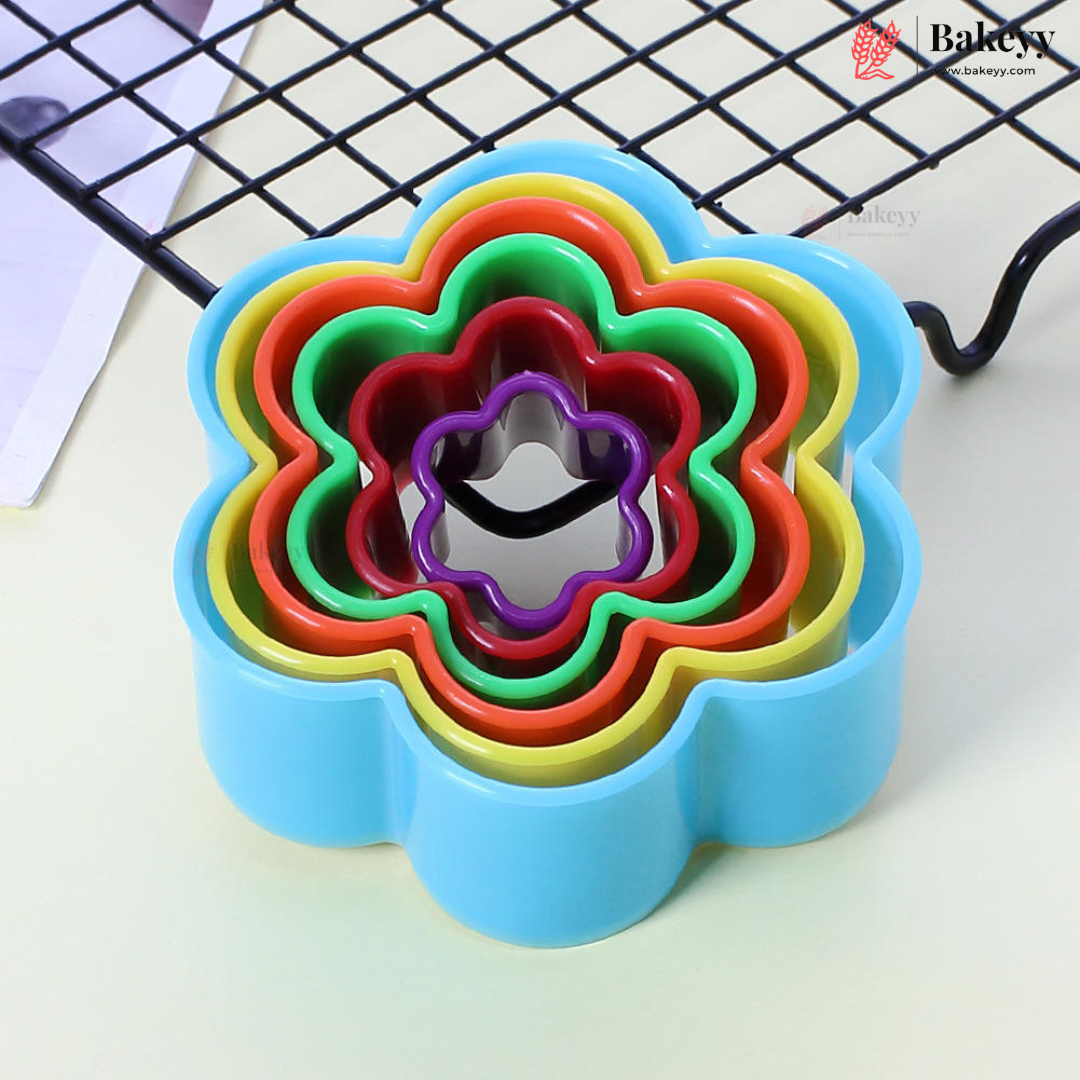 A set of nested flower-shaped cookie cutters in rainbow colors, including blue, red, green, yellow, and purple, with a metal scoring blade visible in the center. The cutters are arranged on a light surface with a black wire cooling rack in the background.