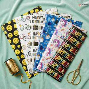 Collection of colorful birthday-themed gift wrap sheets with a spool of ribbon and scissors on a light green fabric background.