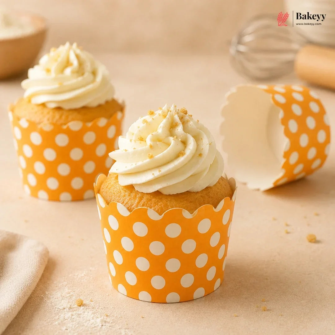 Two cupcakes with white frosting in orange polka dot wrappers on a beige surface, with 'Bakey' branding.