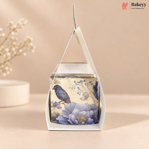 A square white gift box with a clear window and triangular handle, decorated with a vintage-style illustration of a blue bird perched on large lavender-blue flowers against a cream background. The box is photographed on a warm beige surface with soft floral props blurred in the background.