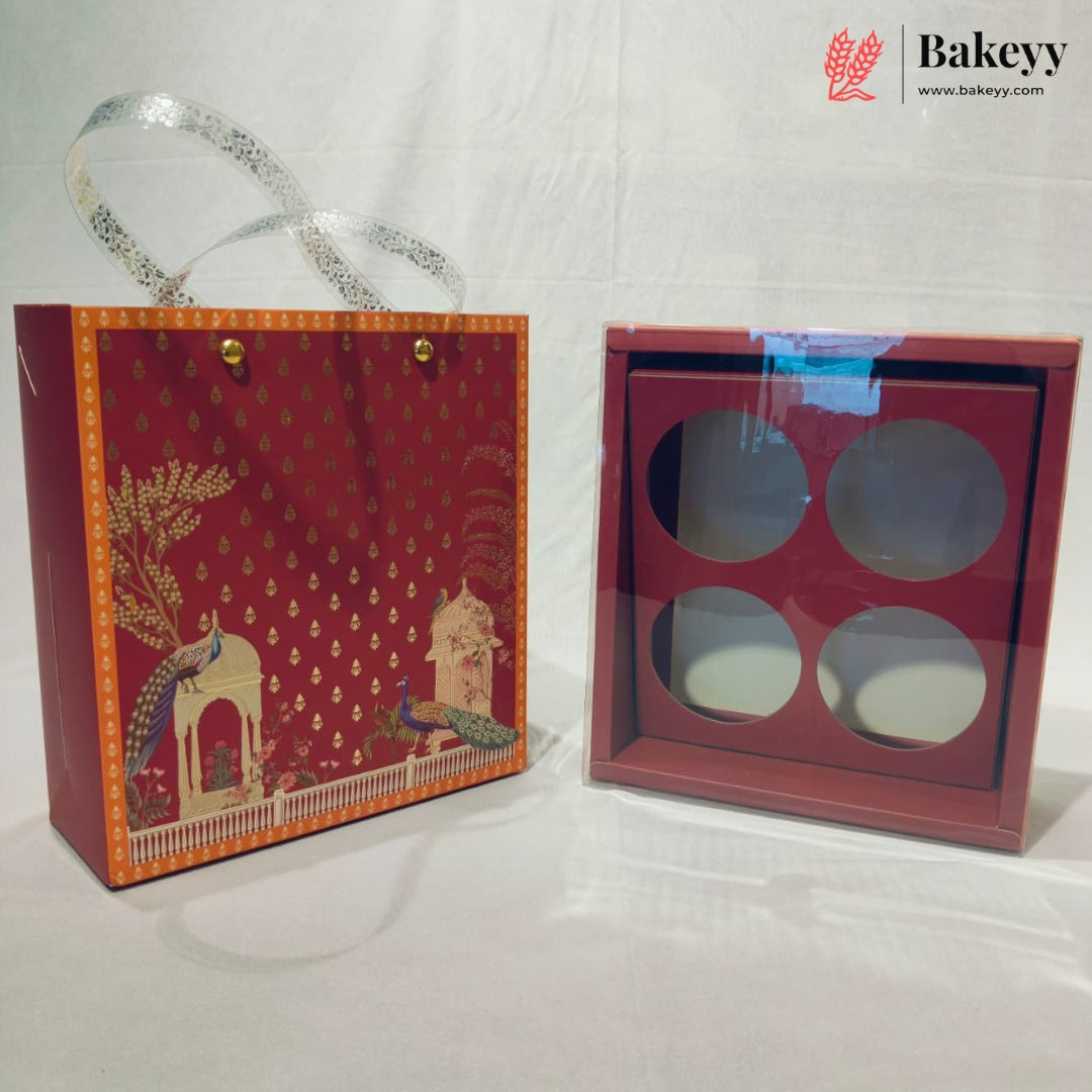 A red gift box set with a decorative printed paper bag featuring peacock and architectural motifs, alongside a matching square box with four circular cutouts and a clear lid, shown on a white fabric background. A Bakeyy logo watermark is visible in the top right.
