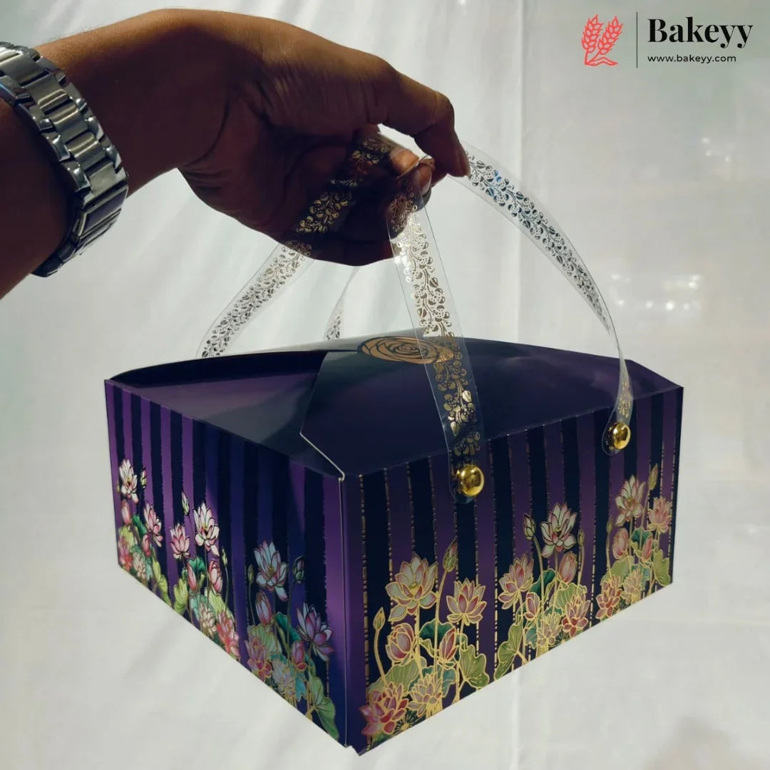 A hand holds a square purple bakery box with floral lotus designs and clear plastic handles. The box features vertical purple stripes and a gold seal on top.