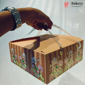 A hand wearing a silver watch holds a decorative cardboard bakery box with floral illustrations and gold-tone hardware against a white backdrop. The Bakeyy logo appears in the top right corner.