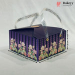 A rectangular bakery gift box with a dark purple lid and floral-patterned sides, shown on a light fabric background with dimension arrows and text indicating 20 cm by 20 cm and 10 cm height. A Bakeyy logo appears in the top right corner.