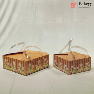 Two decorative rectangular bakery gift boxes with clear plastic handles, featuring a brown striped pattern and a floral border design, displayed on a white fabric backdrop. The Bakeyy logo is visible in the top right corner.