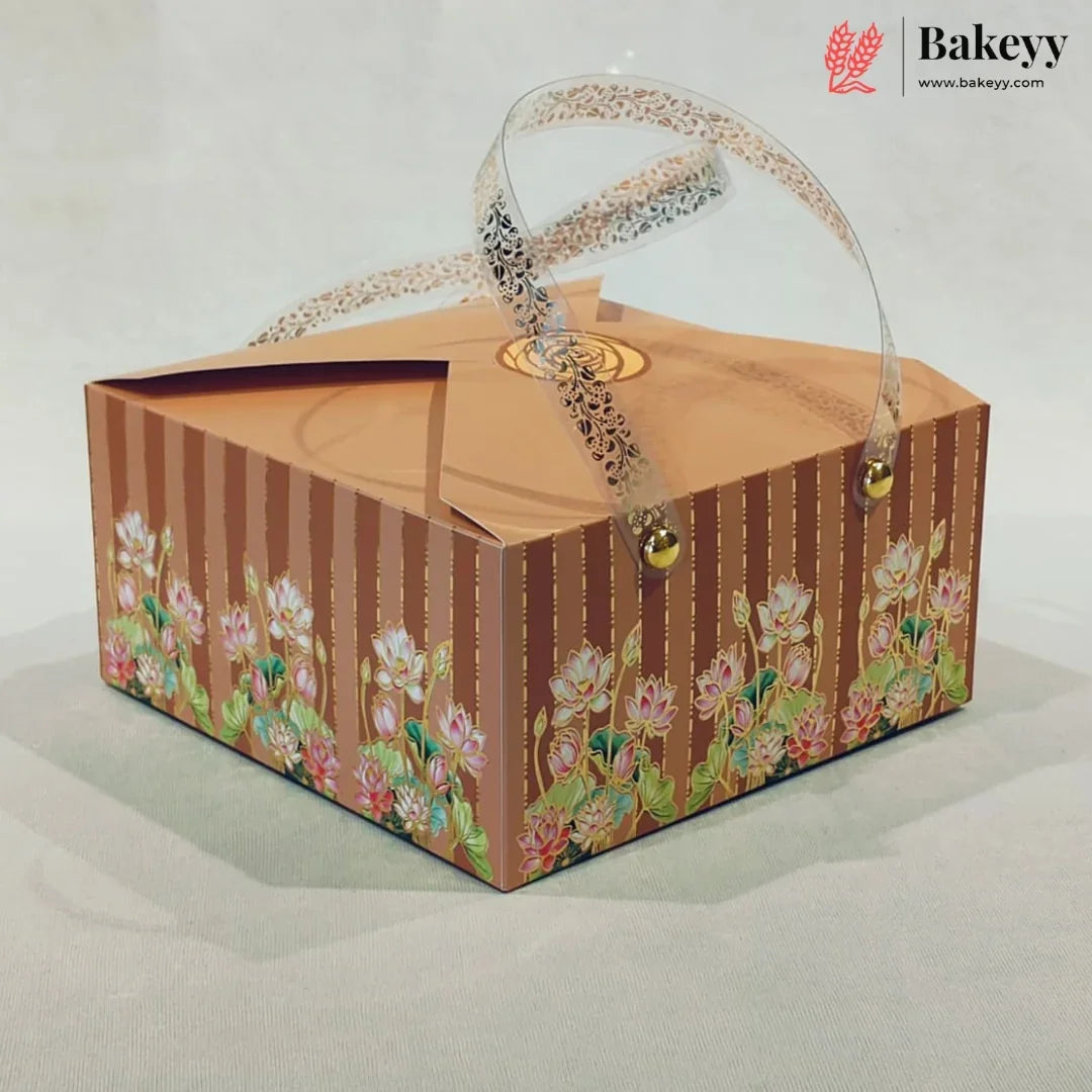 A square bakery gift box with a brown and gold striped pattern, decorated with pink lotus flowers along the bottom. The box features a clear plastic handle with a black lace pattern and small gold bells at the ends, set against a neutral light gray background.