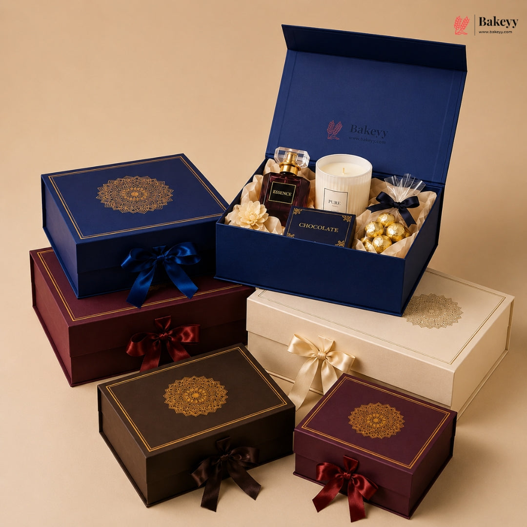 A curated gift box arrangement on a warm beige studio background. An open navy blue Bakeey gift box is open to reveal a candle, chocolates, and small gift items, while several closed gift boxes in navy, burgundy, brown, and cream are arranged around it.