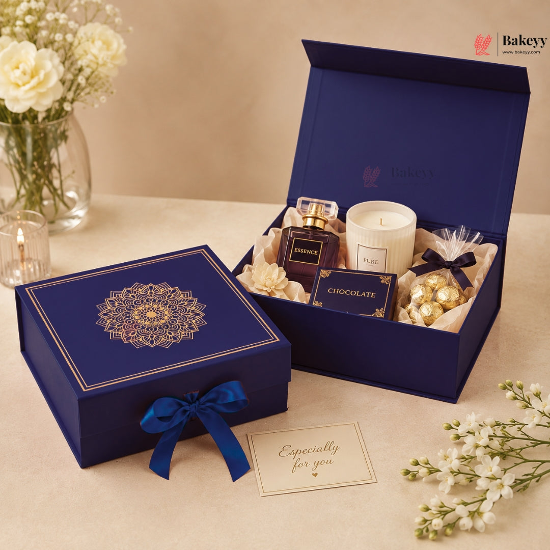 A luxury gift set from Bakey's displayed on a beige table. The set includes a navy blue box with a gold mandala design and ribbon, an open matching box containing a perfume bottle, a white candle, a chocolate box, and wrapped chocolates, along with a 'Especially for you' card and decorative white flowers.