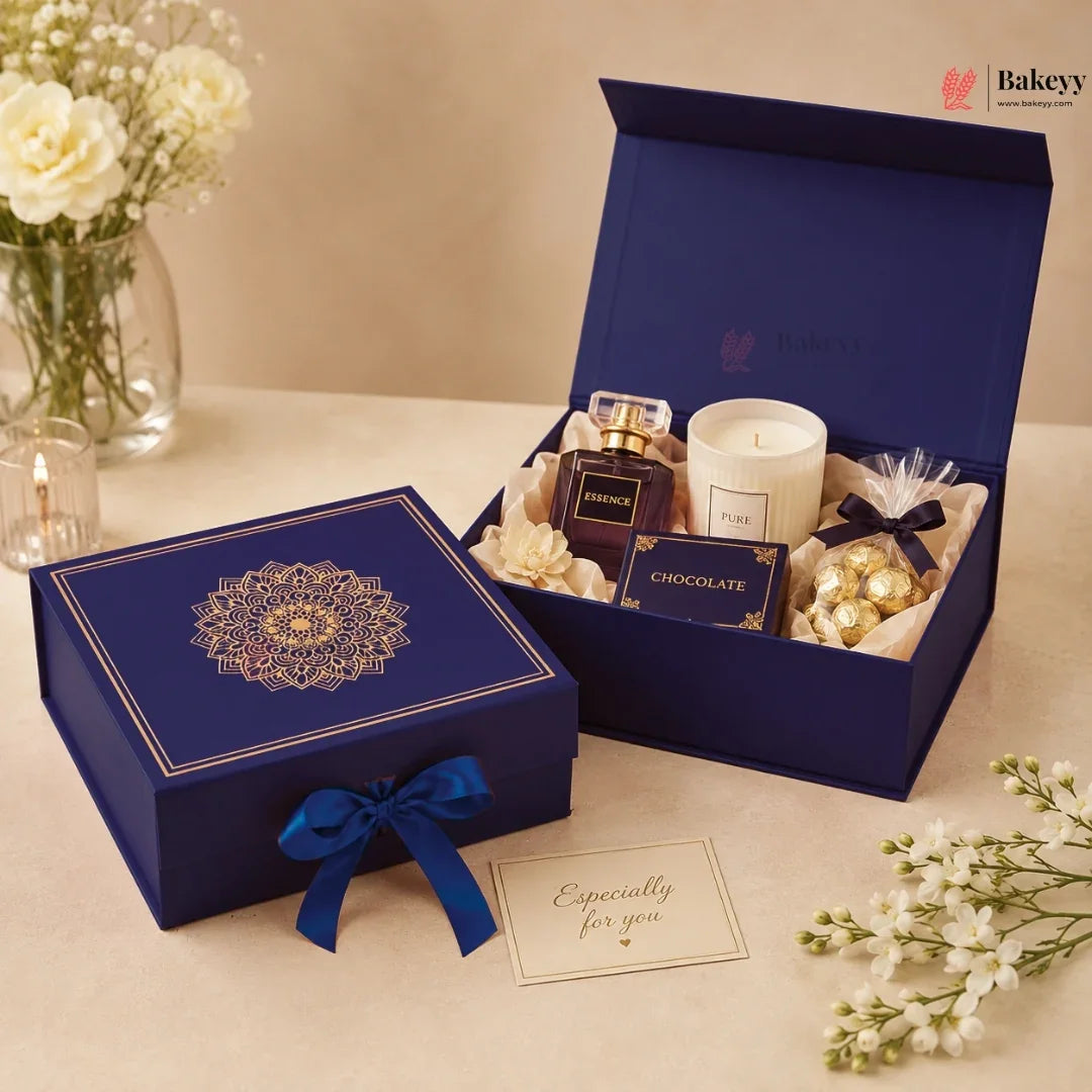 A luxury gift set from Bakey's displayed on a beige table. The set includes a navy blue box with a gold mandala design and ribbon, an open matching box containing a perfume bottle, a white candle, a chocolate box, and wrapped chocolates, along with a 'Especially for you' card and decorative white flowers.