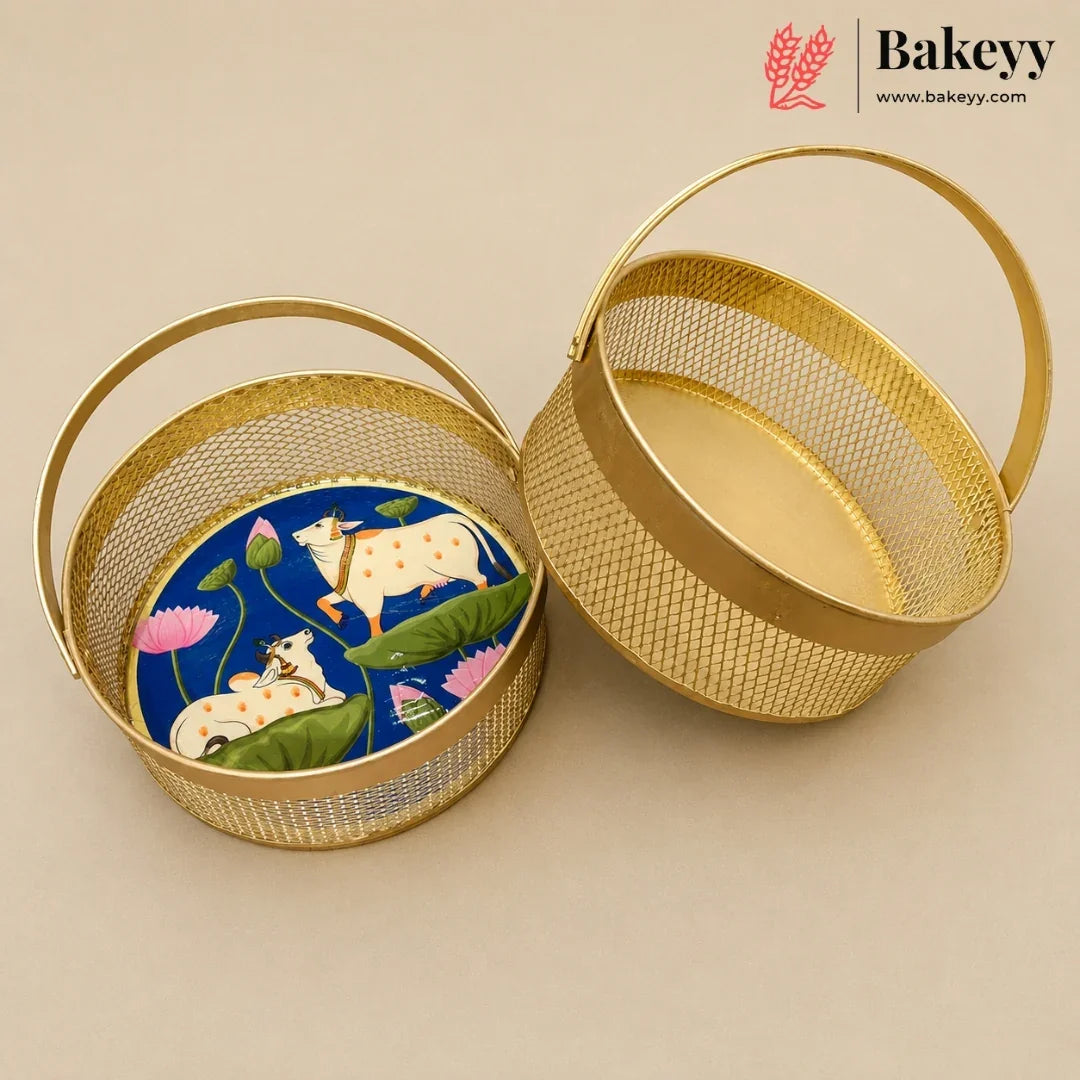 Two round gold metal baskets with mesh sides and top handles, shown on a beige background. One basket is open and displays a colorful printed insert featuring cows, lotus flowers, and green leaves on a blue background.