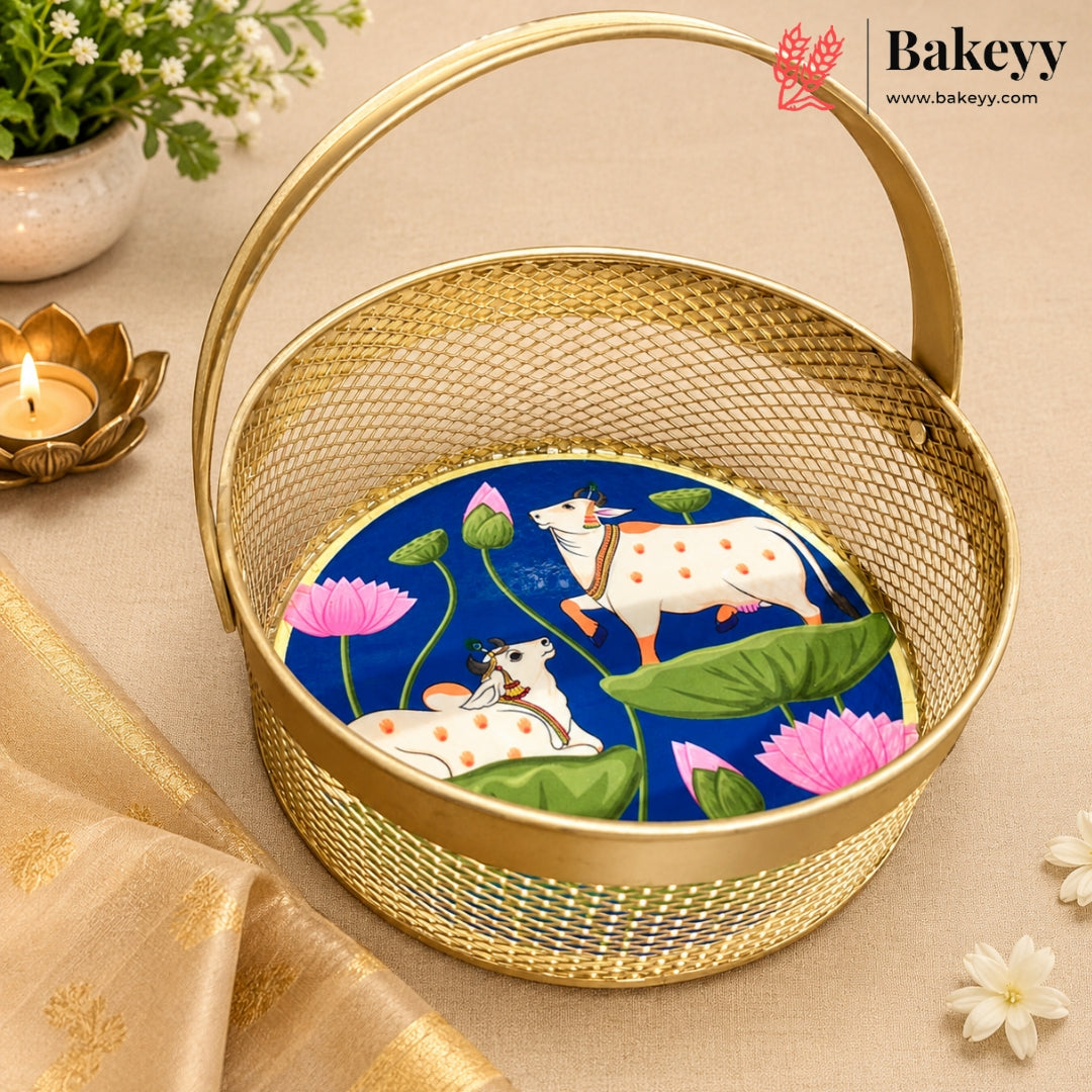 A gold-colored metal wire basket with a handle, featuring a colorful printed bottom insert depicting two white cows with orange spots on a blue background with pink lotus flowers and green leaves. The basket is styled on a beige fabric surface with a gold fabric cloth, a lit candle in a gold lotus holder, and a small potted plant.