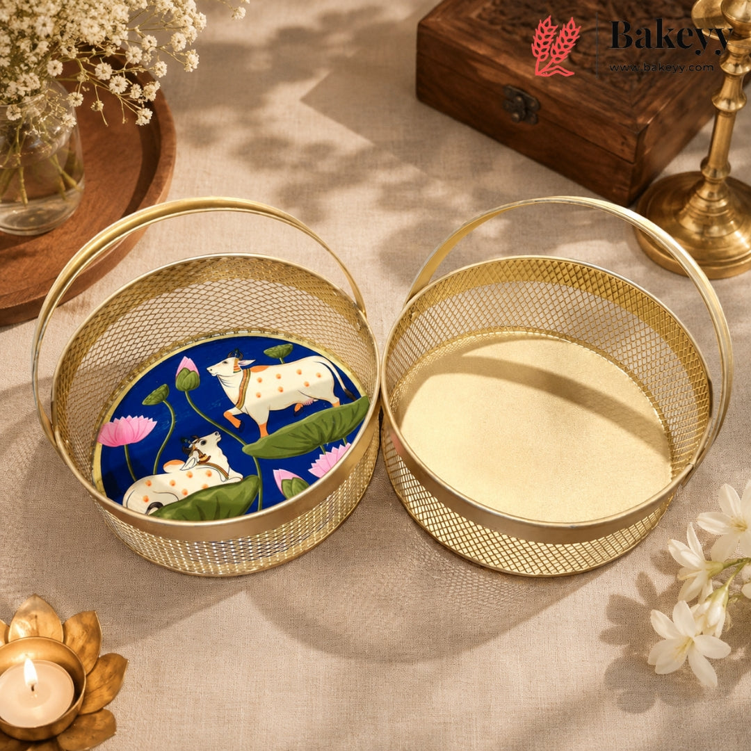 Two round gold wire baskets with handles are displayed on a beige fabric surface. One basket is open and shows a decorative insert with a blue lotus pond design featuring white cows, while the other is open and shows a plain cream-colored interior.