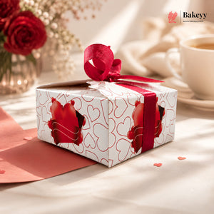 Gift box with red ribbon and heart designs on a table with flowers and a cup in the background, featuring the Bakeyy brand.