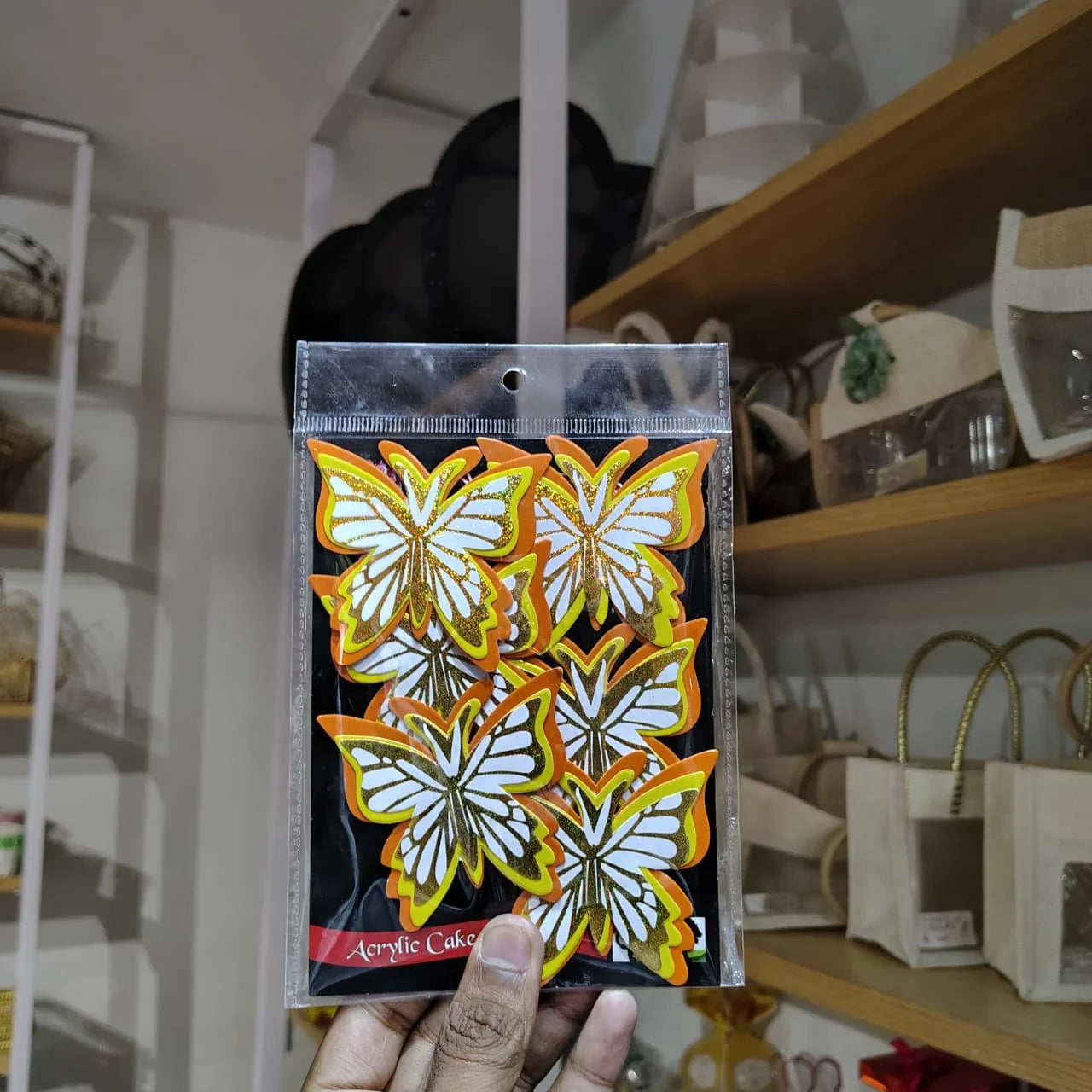 Packaged acrylic cake decorations with butterfly designs held in front of a shelf.