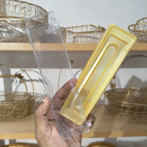 A hand holding a clear plastic rectangular box next to a translucent yellow plastic insert tray, with wire baskets on wooden shelves in the background.