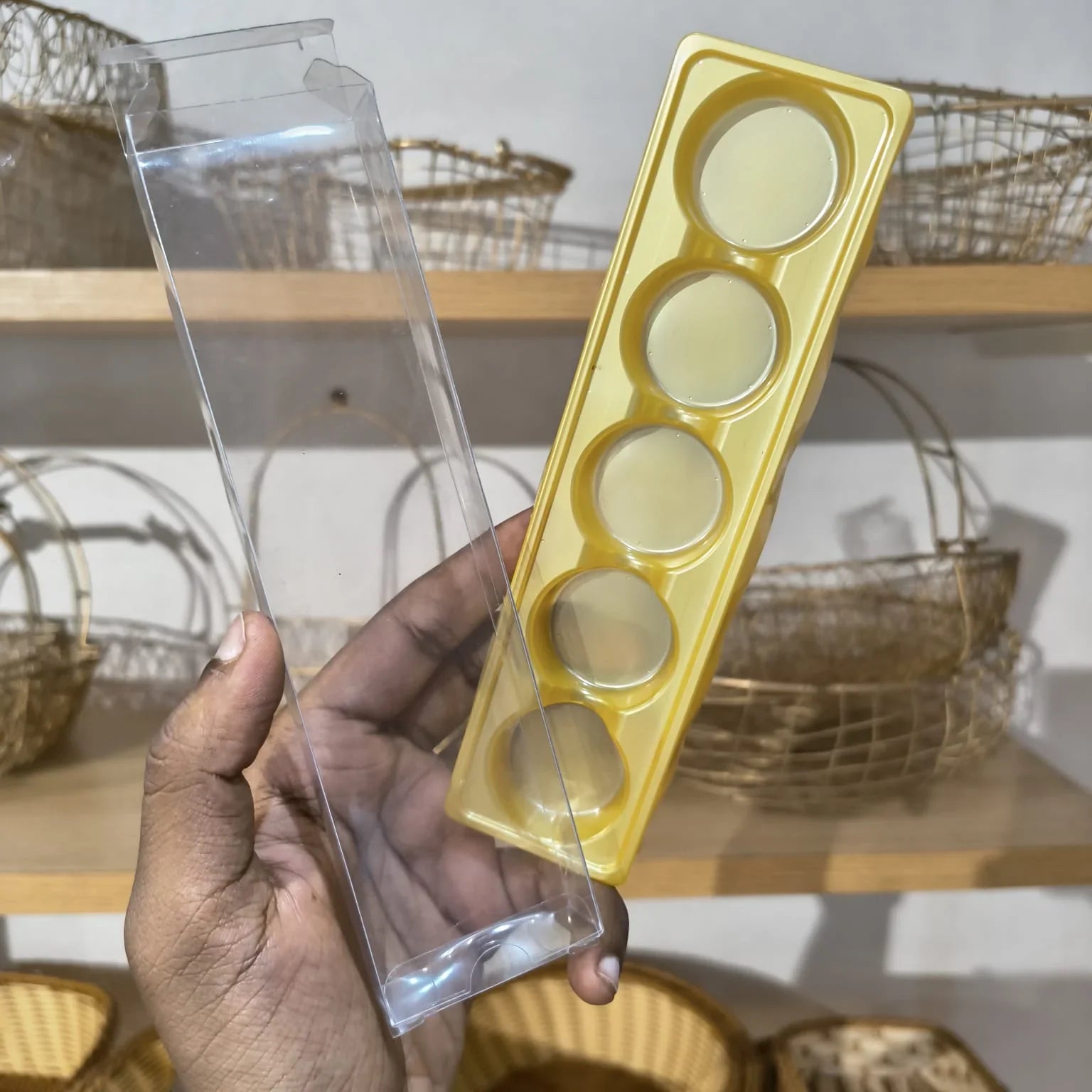 A hand holds a yellow plastic mold tray with five circular cavities next to a clear plastic outer packaging sleeve, with wooden shelves and woven baskets in the background.