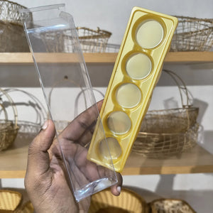 A hand holds a yellow plastic mold tray with five circular cavities next to a clear plastic outer packaging sleeve, with wooden shelves and woven baskets in the background.