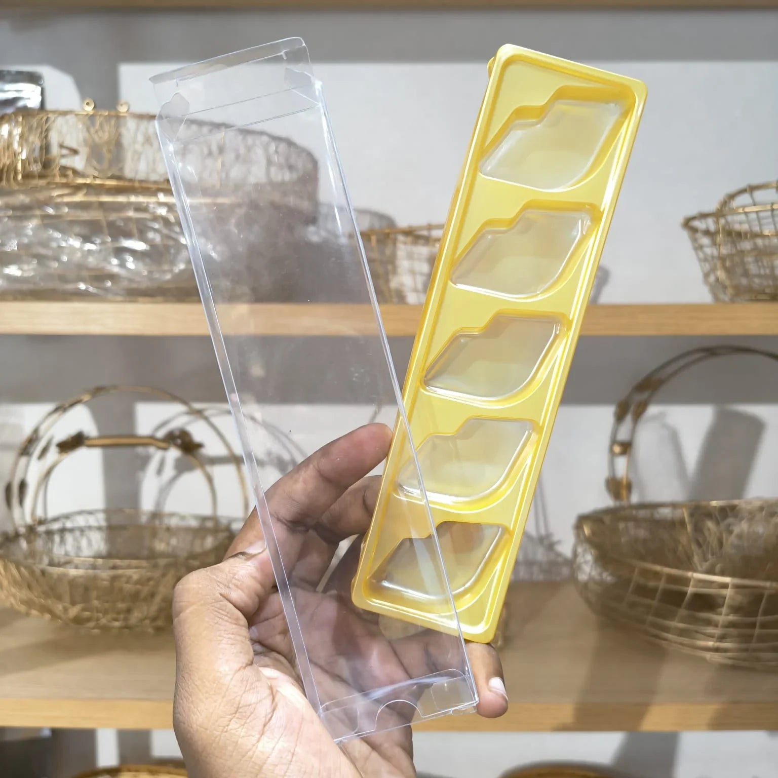 A hand holds a clear plastic rectangular box with four recessed compartments next to a yellow plastic tray with matching compartment shapes, against a blurred background of shelves with decorative baskets.