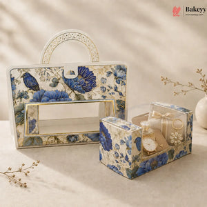 A decorative gift box set with a large rectangular carrying case and a smaller square presentation box, both covered in a blue floral and bird pattern with gold trim, styled on a beige surface with dried flowers and soft shadows.