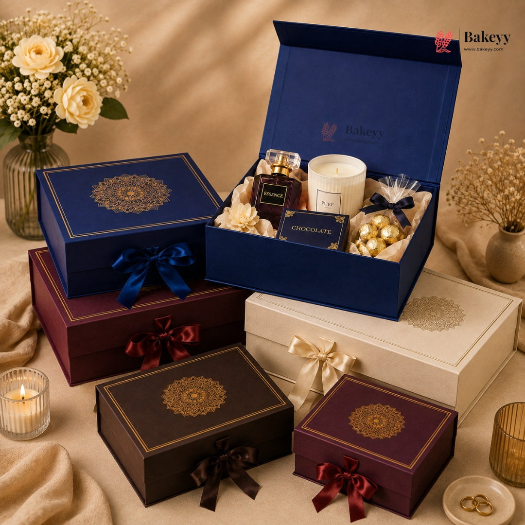 A styled gift set display featuring multiple luxury bakery boxes in navy blue, burgundy, and cream, with one open box revealing chocolates, a candle, and a small jar. The scene is decorated with roses, dried flowers, and candles on a beige fabric background.