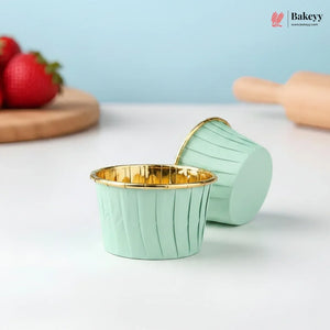 Mint green cupcake liners with gold rims on a white surface, with strawberries and a rolling pin in the background.
