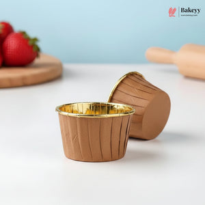 Brown muffin cups with gold interior on a white surface, with strawberries and a rolling pin in the background.