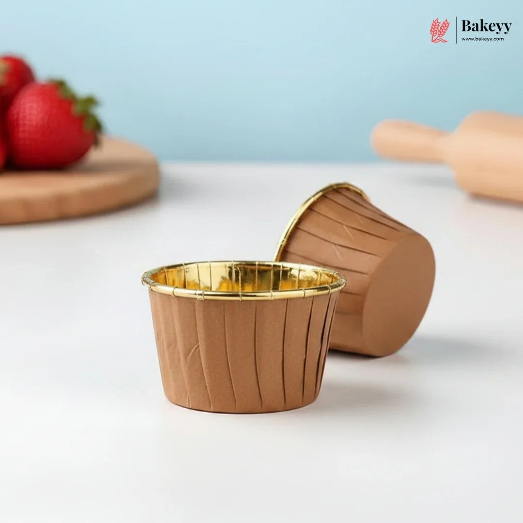 Brown muffin cups with gold interior on a white surface, with strawberries and a rolling pin in the background.