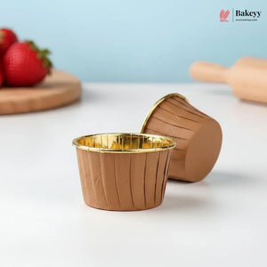 Brown muffin cups with gold interior on a white surface, with strawberries and a rolling pin in the background.