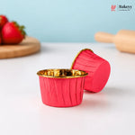 Red silicone muffin cups with gold interior on a light surface, with strawberries and a rolling pin in the background.