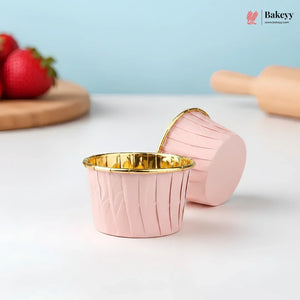 Pink cupcake liners with gold interior on a light surface, with strawberries and a rolling pin in the background.