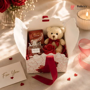 Gift box with teddy bear, Lindor chocolate, and perfume on a soft surface with 'Bakeyy' branding.