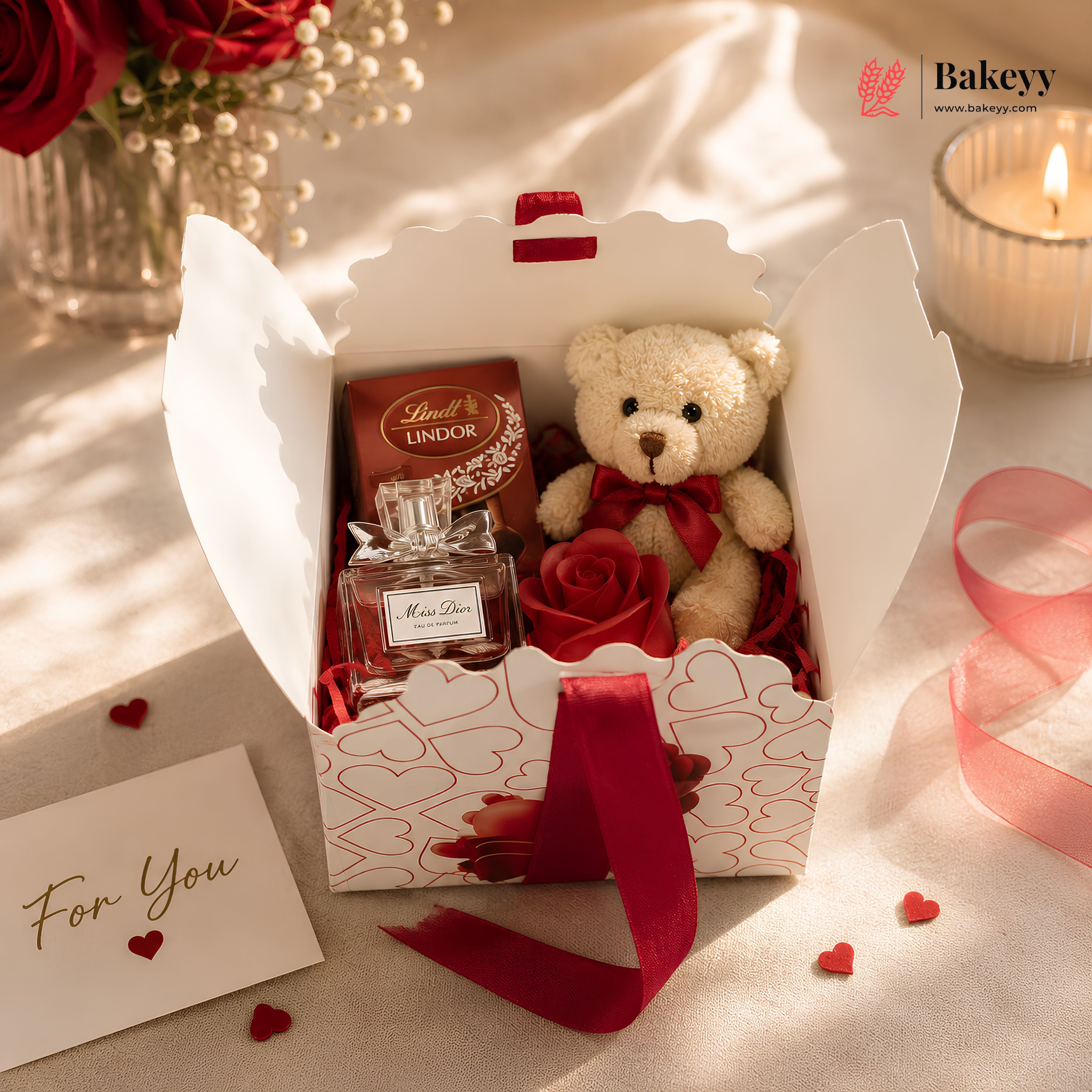 Gift box with teddy bear, Lindor chocolate, and perfume on a soft surface with 'Bakeyy' branding.