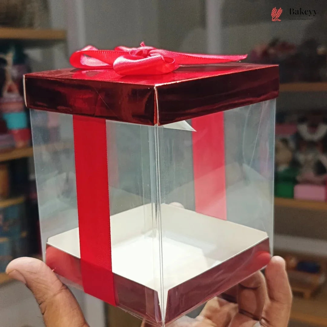 Clear gift box with red ribbon held by a hand, blurred indoor background