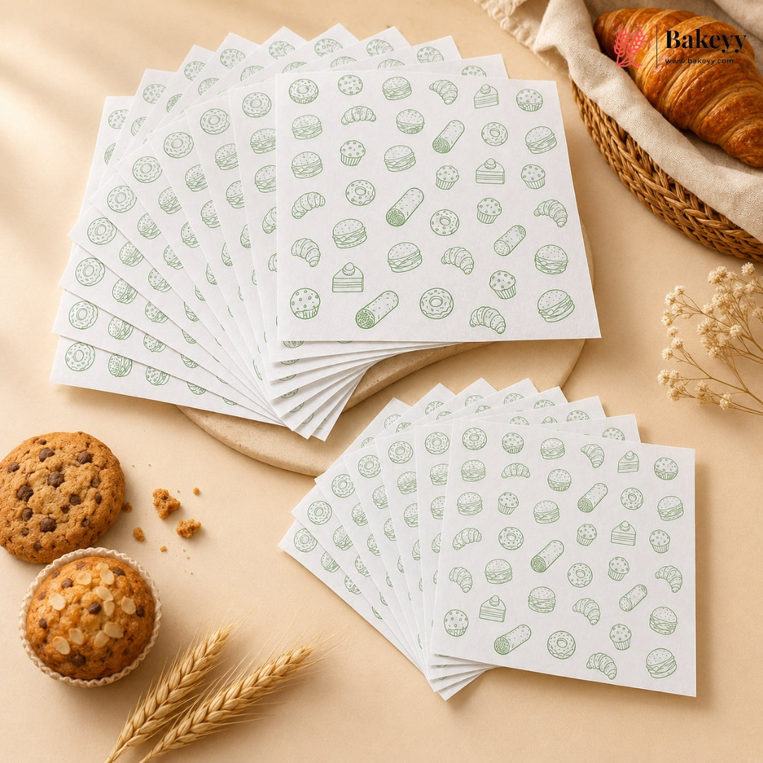 Two stacks of white baking mats or napkins printed with a light green pattern of various baked goods, cookies, pastries, and bread, arranged on a beige surface with decorative food props like muffins, a croissant, wheat, and dried flowers. The brand name 'Bakeyy' is printed on the top stack.