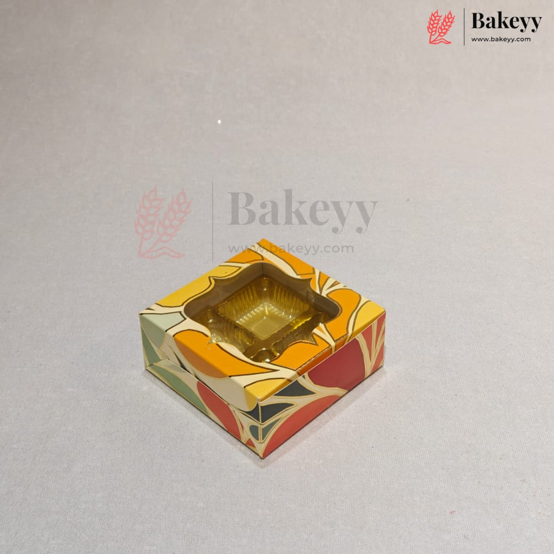 A square decorative bakery box with a clear plastic window on top, showing a gold-colored tray inside. The box features a colorful abstract pattern in orange, yellow, red, and green on a light gray background with Bakeey watermarks.
