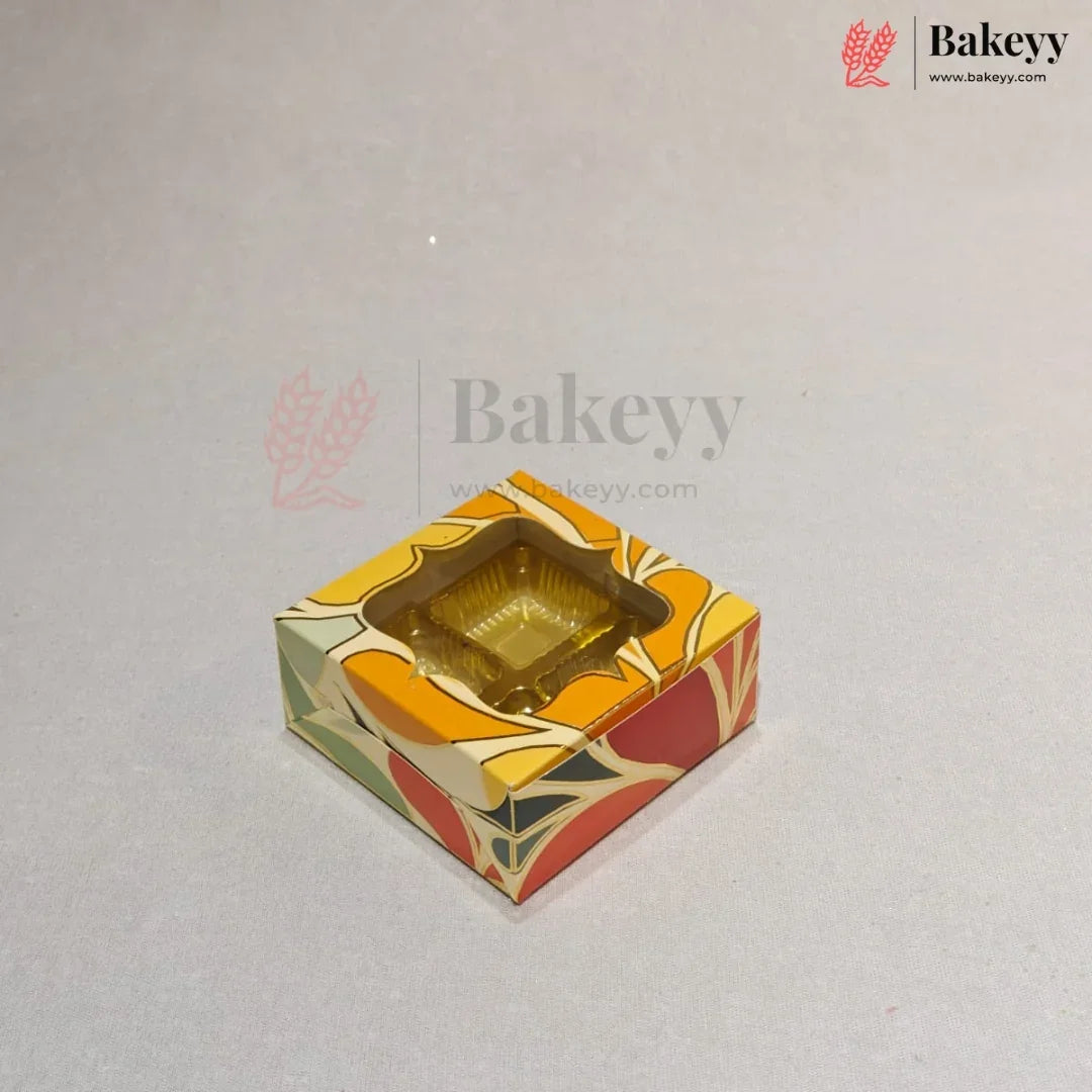 A square decorative bakery box with a clear plastic window on top, showing a gold-colored tray inside. The box features a colorful abstract pattern in orange, yellow, red, and green on a light gray background with Bakeey watermarks.