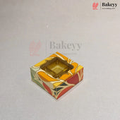 A square decorative bakery box with a clear plastic window on top, showing a gold-colored tray inside. The box features a colorful abstract pattern in orange, yellow, red, and green on a light gray background with Bakeey watermarks.
