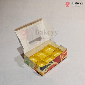 An open bakery box with a hinged lid, featuring a gold foil crumb tray inside and a printed multicolor pattern on the exterior. The Bakeyy logo is visible in the top right corner.