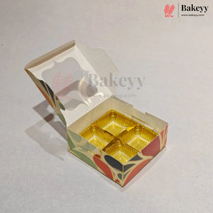 An open custom cardboard bakery box with four compartments lined with shiny gold foil, featuring a cream base with red, green, and black floral patterns. The Bakeyy logo is visible in the top right corner.