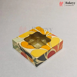 A square, multi-compartment gift box with a colorful retro floral pattern in yellow, orange, red, and green on a cream background, featuring a clear window on top. The Bakeyy brand logo is visible in the top right corner.