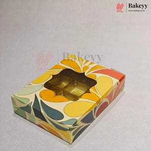 A square bakery box with a colorful retro floral pattern in yellow, orange, green, and red on a cream background, featuring a clear window showing a gold-colored tray inside. The Bakeyy logo is visible in the top right corner.