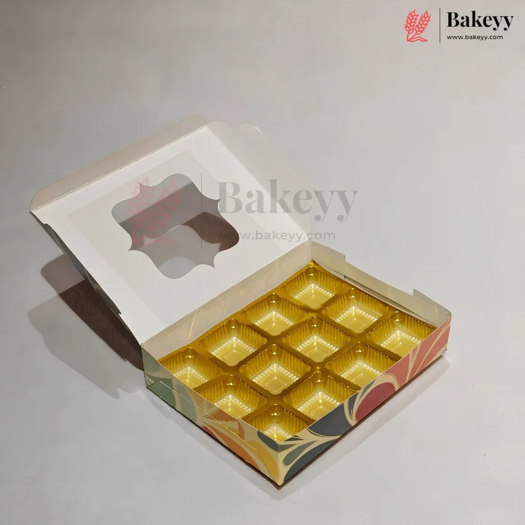 An open bakery box with a hinged lid and a clear window, showing 12 individual gold-colored cupcake or muffin liners arranged inside. The box has a white outer flap and a colorful printed base with green, orange, and red patterns.