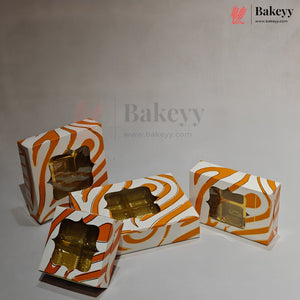 Four decorative bakery boxes with a white and orange wavy pattern and clear windows showing gold foil-wrapped pastries inside, arranged on a light gray studio surface.