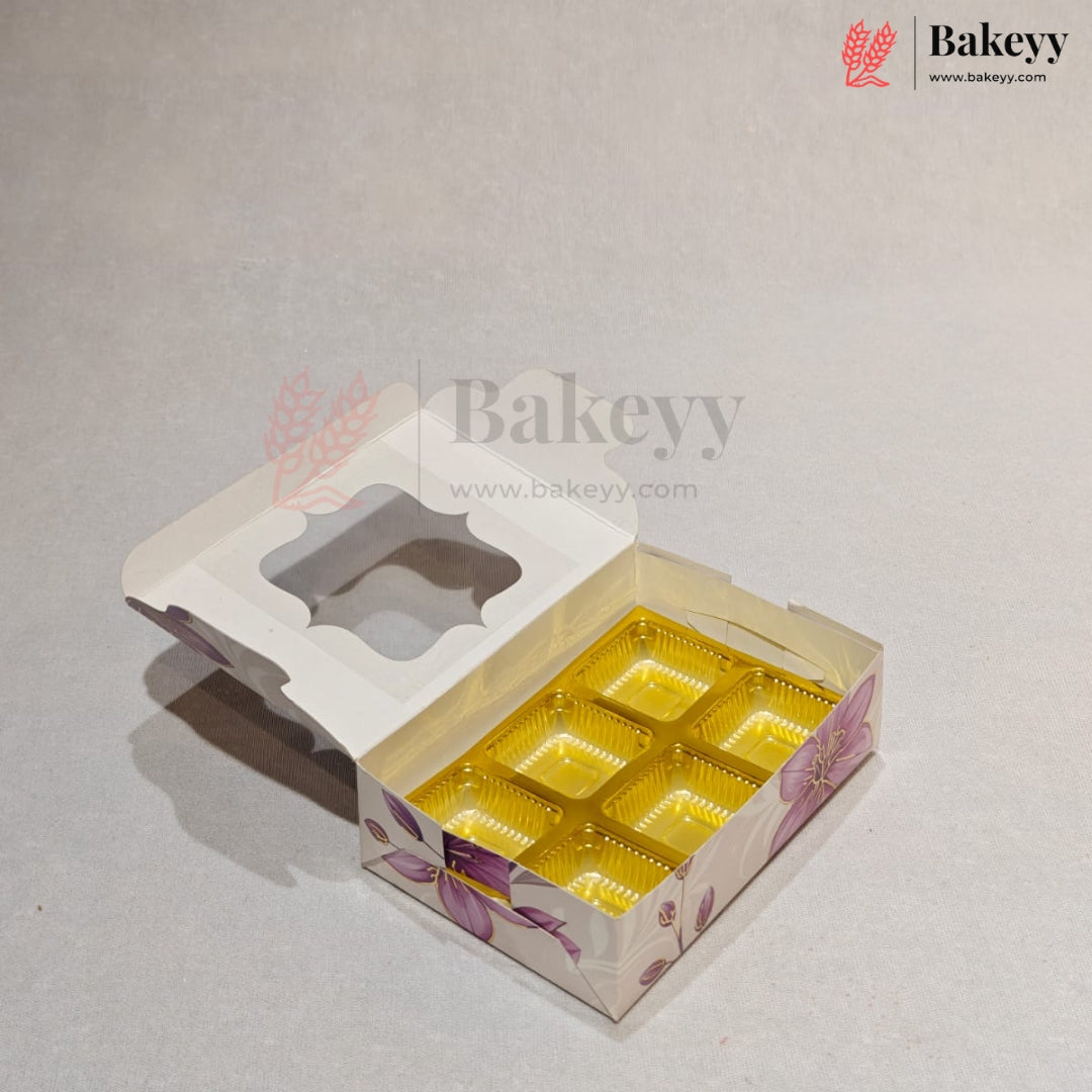 A bakery box with a hinged lid, featuring a floral pattern in purple and pink on a white base, with six gold foil compartments inside. The box is shown open on a light gray background with Bakeyy branding.