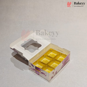 A bakery box with a hinged lid, featuring a floral pattern in purple and pink on a white base, with six gold foil compartments inside. The box is shown open on a light gray background with Bakeyy branding.