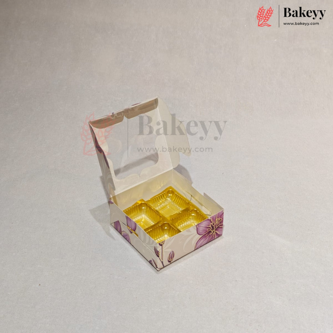 A small open bakery box with a clear window and a yellow tray inside, decorated with a purple floral pattern on a light beige background. The Bakeyy logo is visible in the top right corner.