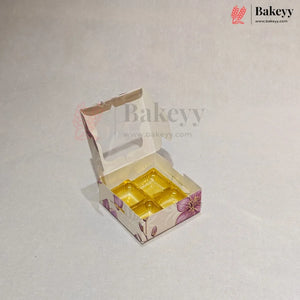 A small open bakery box with a clear window and a yellow tray inside, decorated with a purple floral pattern on a light beige background. The Bakeyy logo is visible in the top right corner.