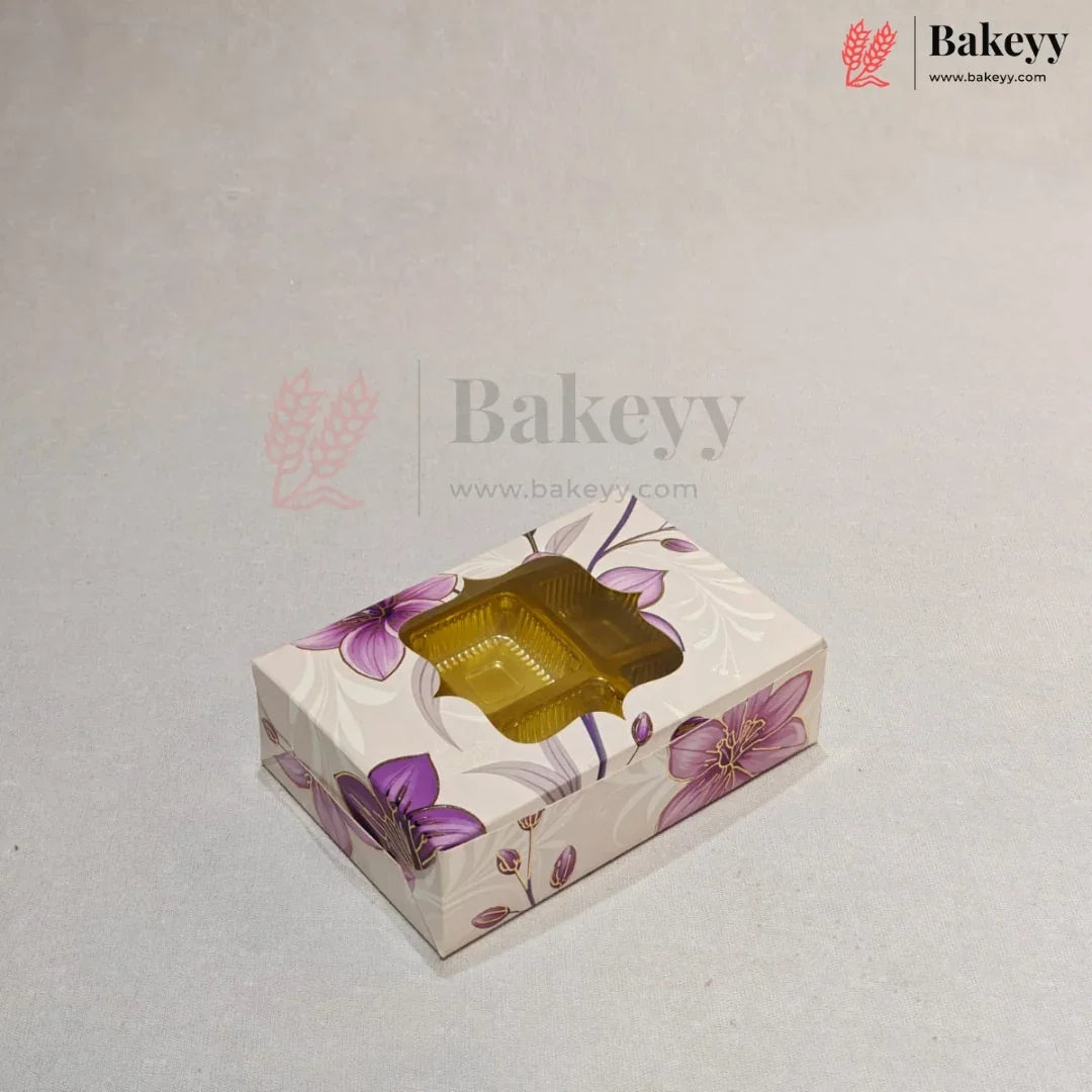 A rectangular bakery box with a floral pattern in purple and pink on a cream background, featuring a clear plastic window on top showing a golden-brown pastry inside. The image has a watermark for 'Bakeyy' on a light gray background.