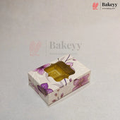 A rectangular bakery box with a floral pattern in purple and pink on a cream background, featuring a clear plastic window on top showing a golden-brown pastry inside. The image has a watermark for 'Bakeyy' on a light gray background.