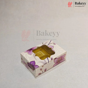 A rectangular bakery box with a floral pattern in purple and pink on a cream background, featuring a clear plastic window on top showing a golden-brown pastry inside. The image has a watermark for 'Bakeyy' on a light gray background.